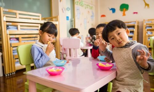 Kids seated around a table in a colorful classroom, eating snacks happily.
